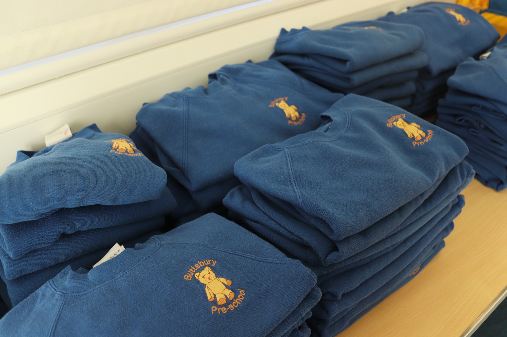 A pile of navy blue jumpers with the Buttsbury Pre-school logo which is a light brown bear.