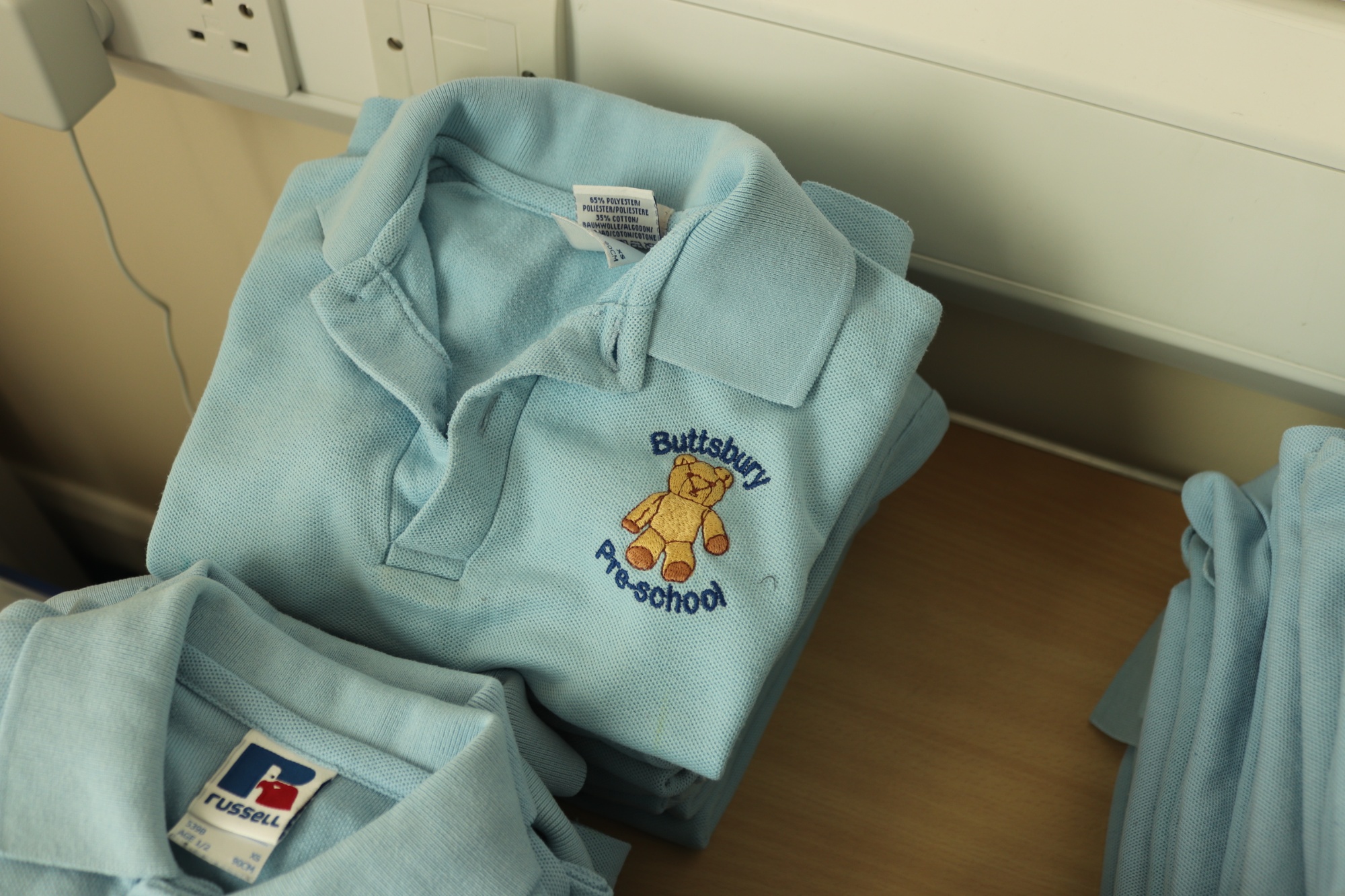 A pile of light blue t-shirts with the Buttsbury Pre-school logo which is a light brown bear.